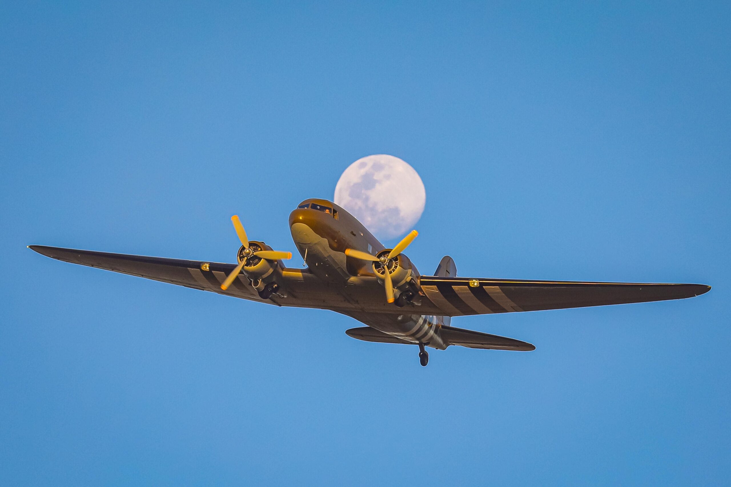 Douglas C-47 Dakota in wartime camouflage with invasion stripes, flying against a blue sky with the moon behind it.
